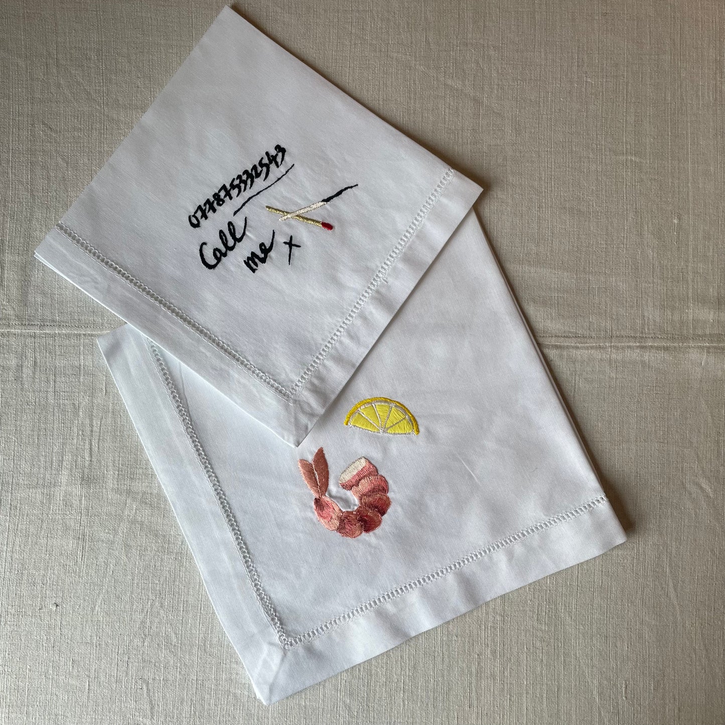 White napkin with the words 'call me', a collection of numbers and a two matchsticks embroidered on. Underneath is another white napkin with an embroidered prawn and lemon on