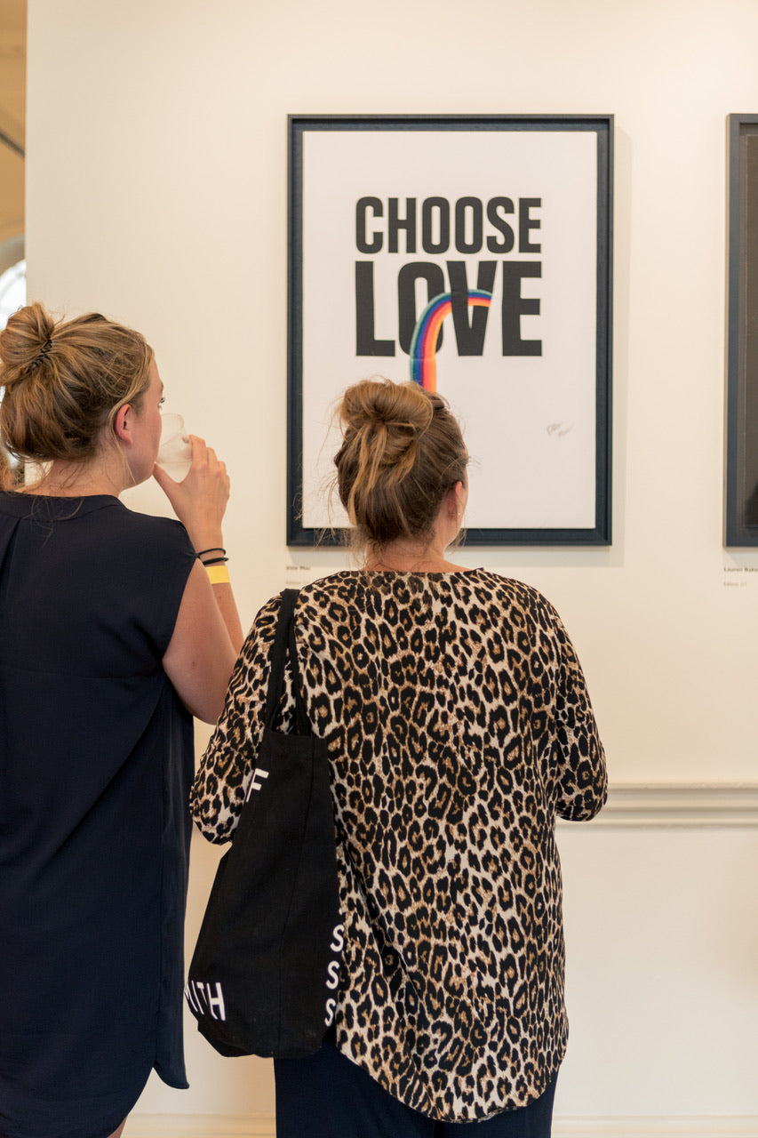 The backs of two people, one in a black top and one in a leopard print top are looking at the Choose Love artwork with embroidered rainbow. The artwork is in a black frame on a white wall