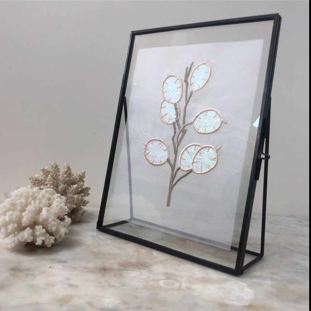 Sheer silk organza honesty embroidery seen in a black metal and glass picture frame on a grey marble surface with two bits of coral to the left of the picture frame