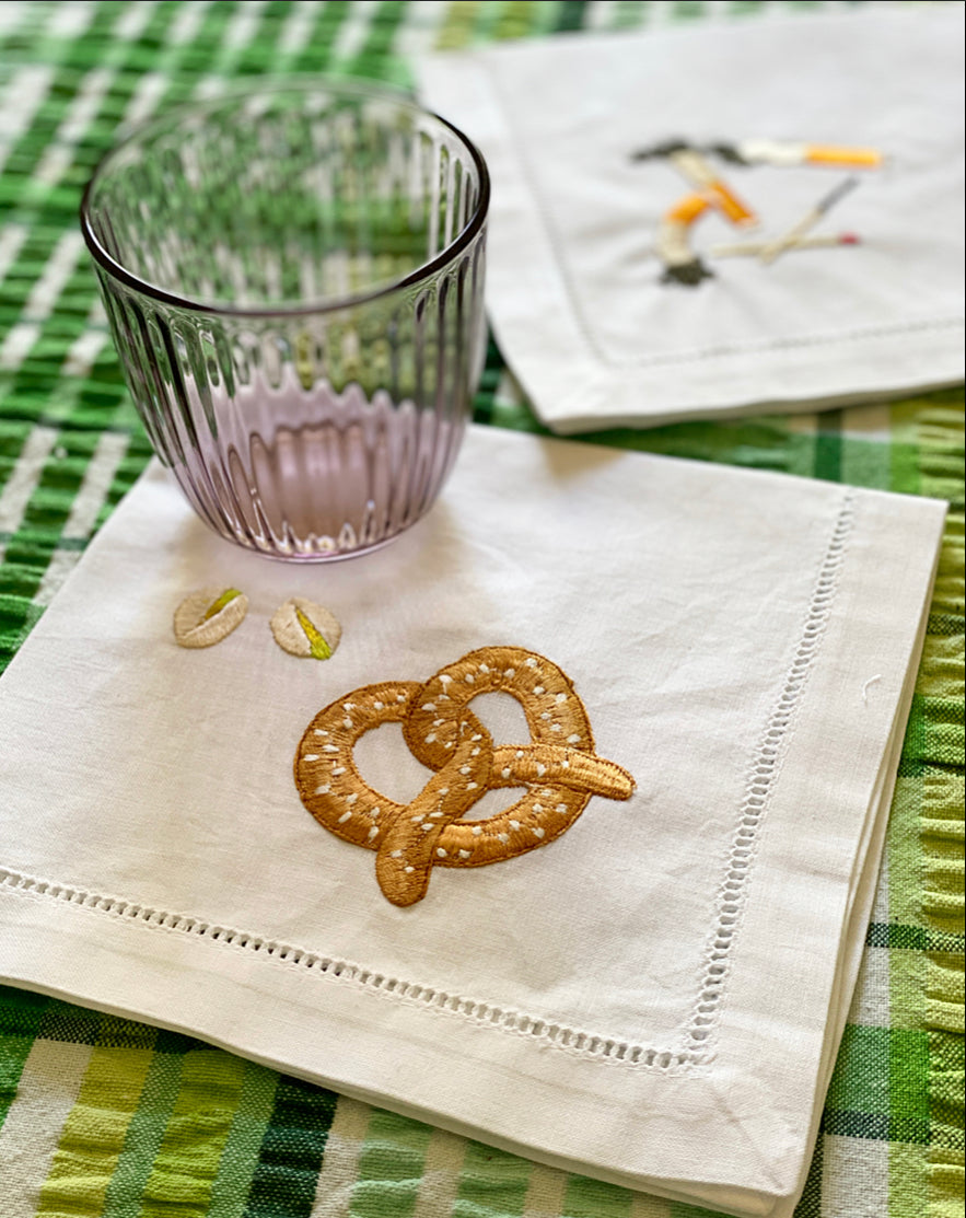 White napkin with embroidered pretzel and pistachios on with a glass on the corner and both items placed ona checkered green tablecloth
