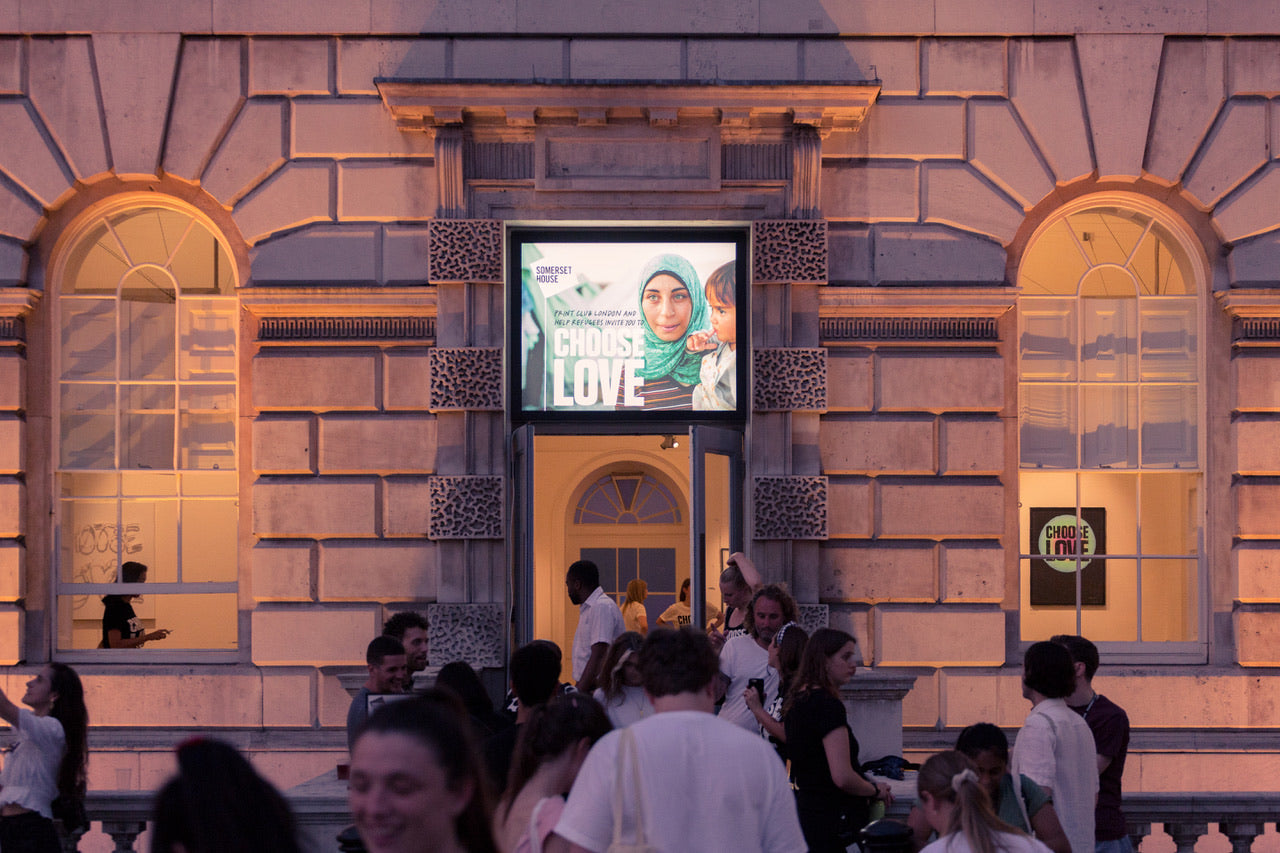 Choose Love exhibition seen from outside of building with open front door and two arched windows where other artwork is visible, a crowd of happy faces is seen in front of the building