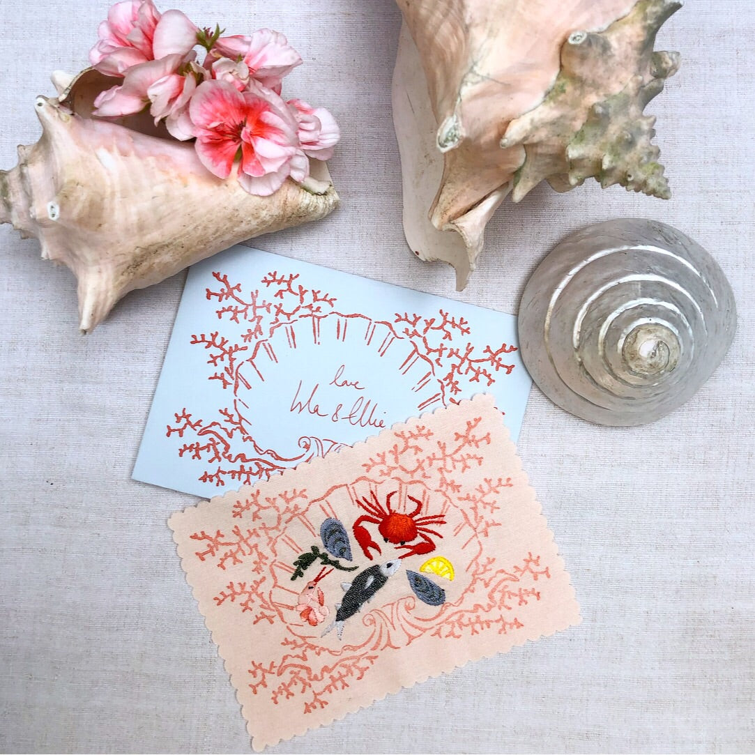 'Fruits de Mer' embroidered piece and white postcard which says 'love Isla & Ellie' and three shells are posed on a white cloth