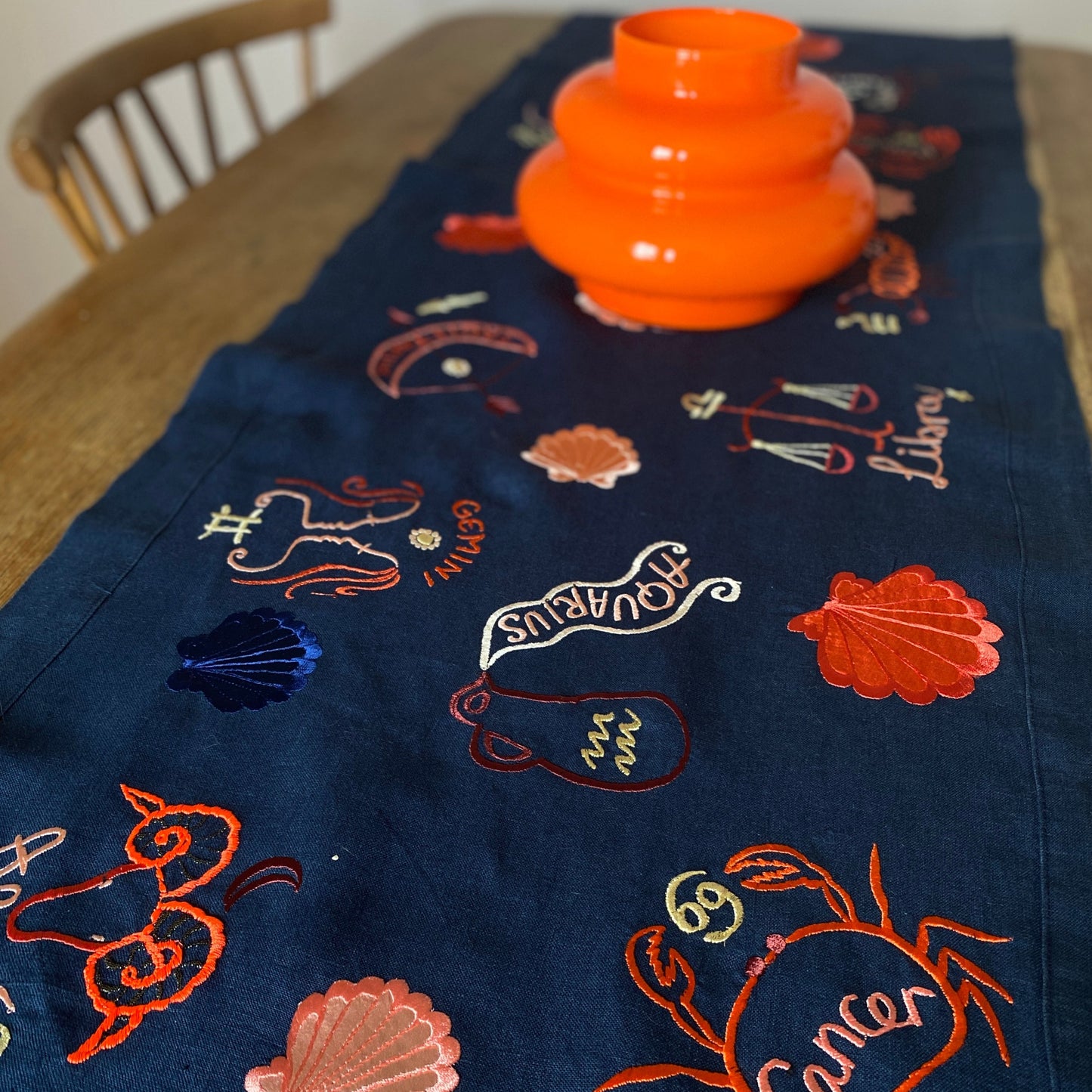 Zodiac embroidered artwork banner laid on a wooden table with orange vase sat on top of the banner