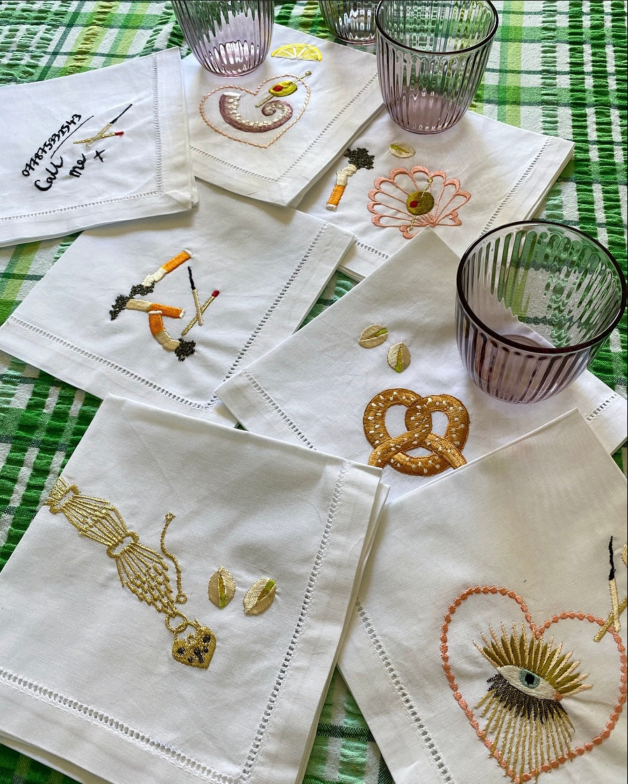 Collection of Alma Berrow X Ellie Mac Embroidery collaborative napkins arranged on table with a green check table cloth and a few drinking glasses