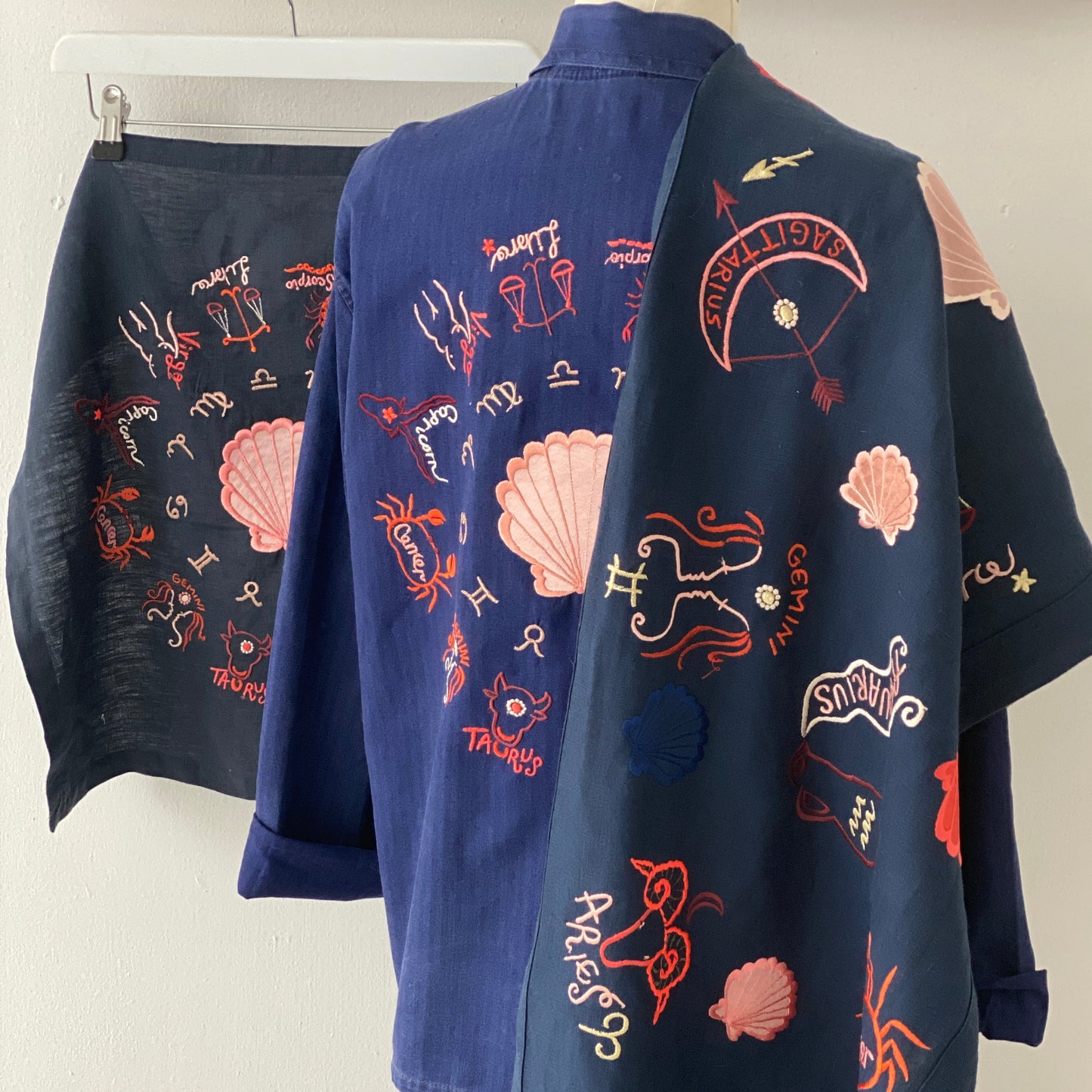 Zodiac artwork in banner shape draped over blue jacket that has more zodiac signs embroidered on the back of the jacket. The square zodiac embroidered wheel can also be seen in the background, behind the jacket