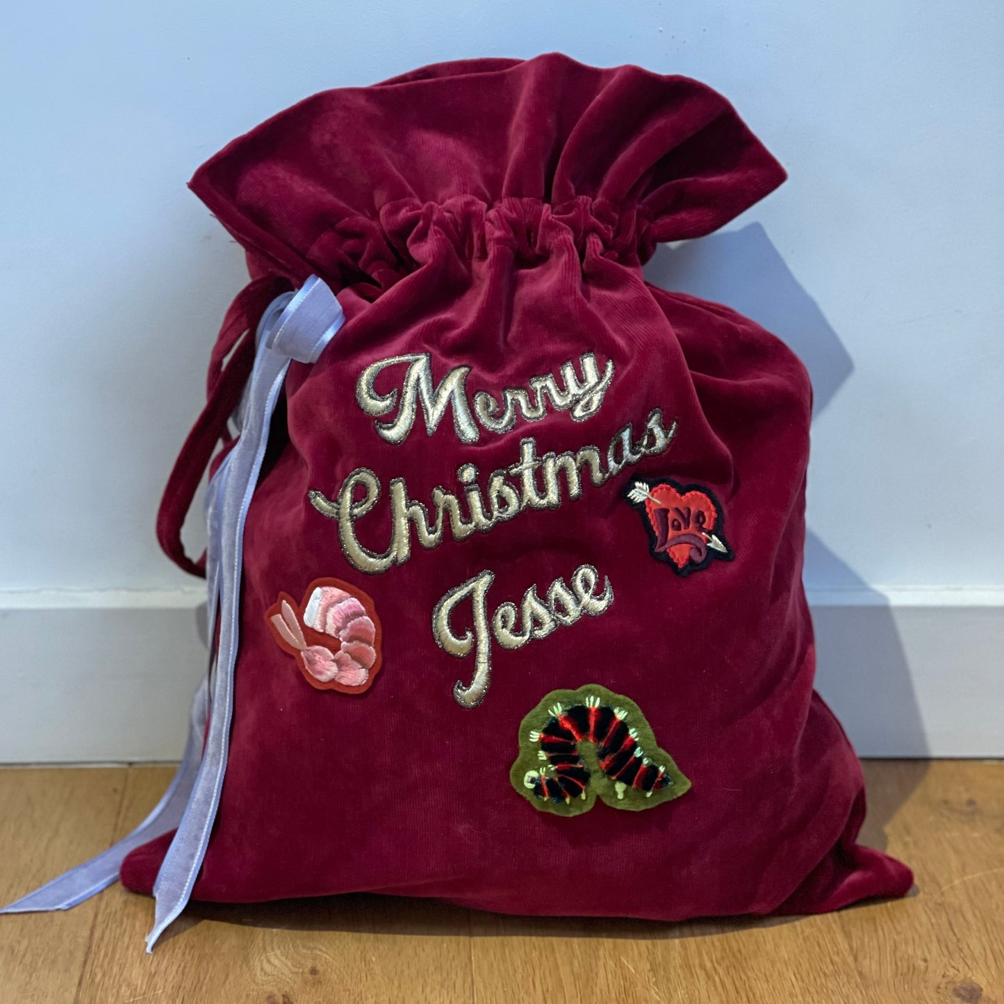 Maroon velvet large drawstring sack against pale blue painted wall. The sack has Merry Christmas Jesse embroidered in gold thread and three patches, a caterpilla, love heart and the prawn tail