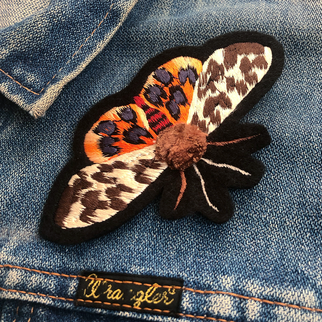 Close-up pom-pom tiger moth embroidered patch on front shoulder of blue denim jacket