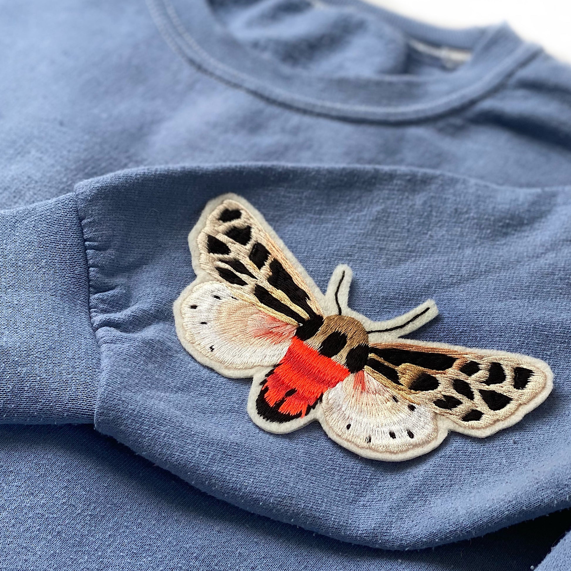 Neon tiger moth embroidered patch show on the sleeve of a light blue jumper