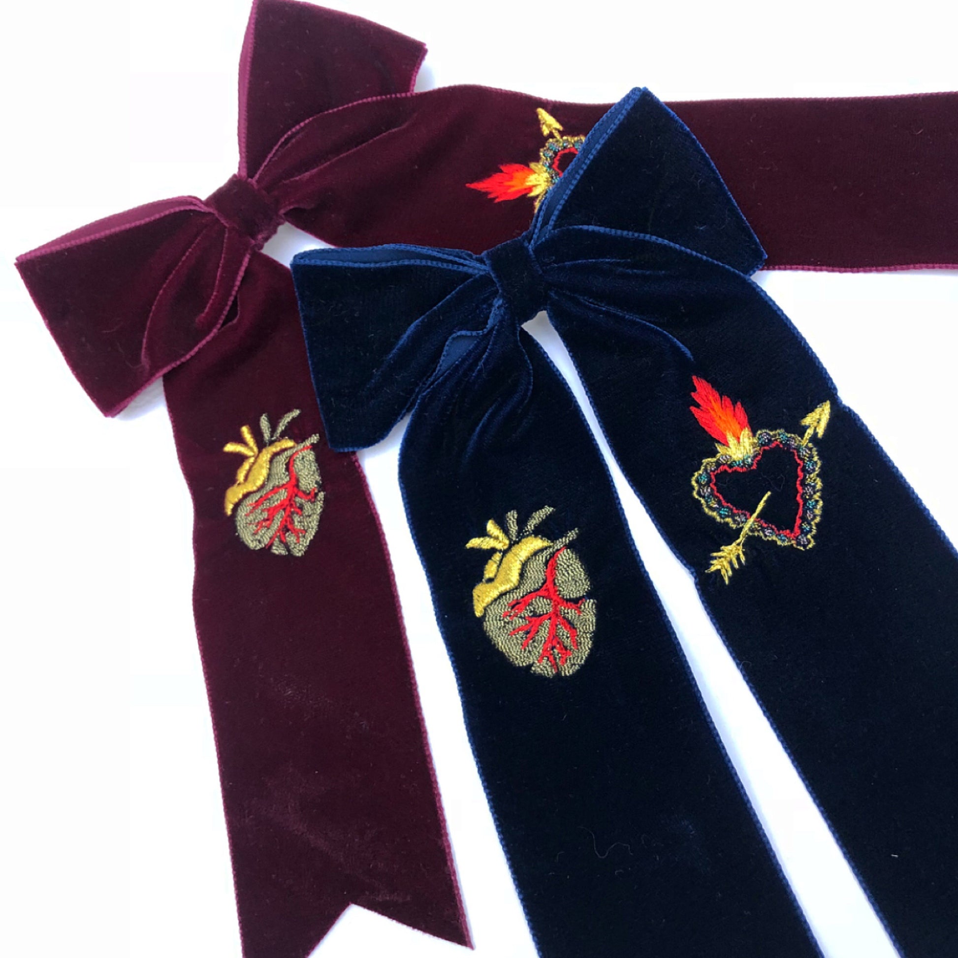 A maroon velvet embroidered hair bow with a navy blue embroidered hair bow lying on top, on a white background