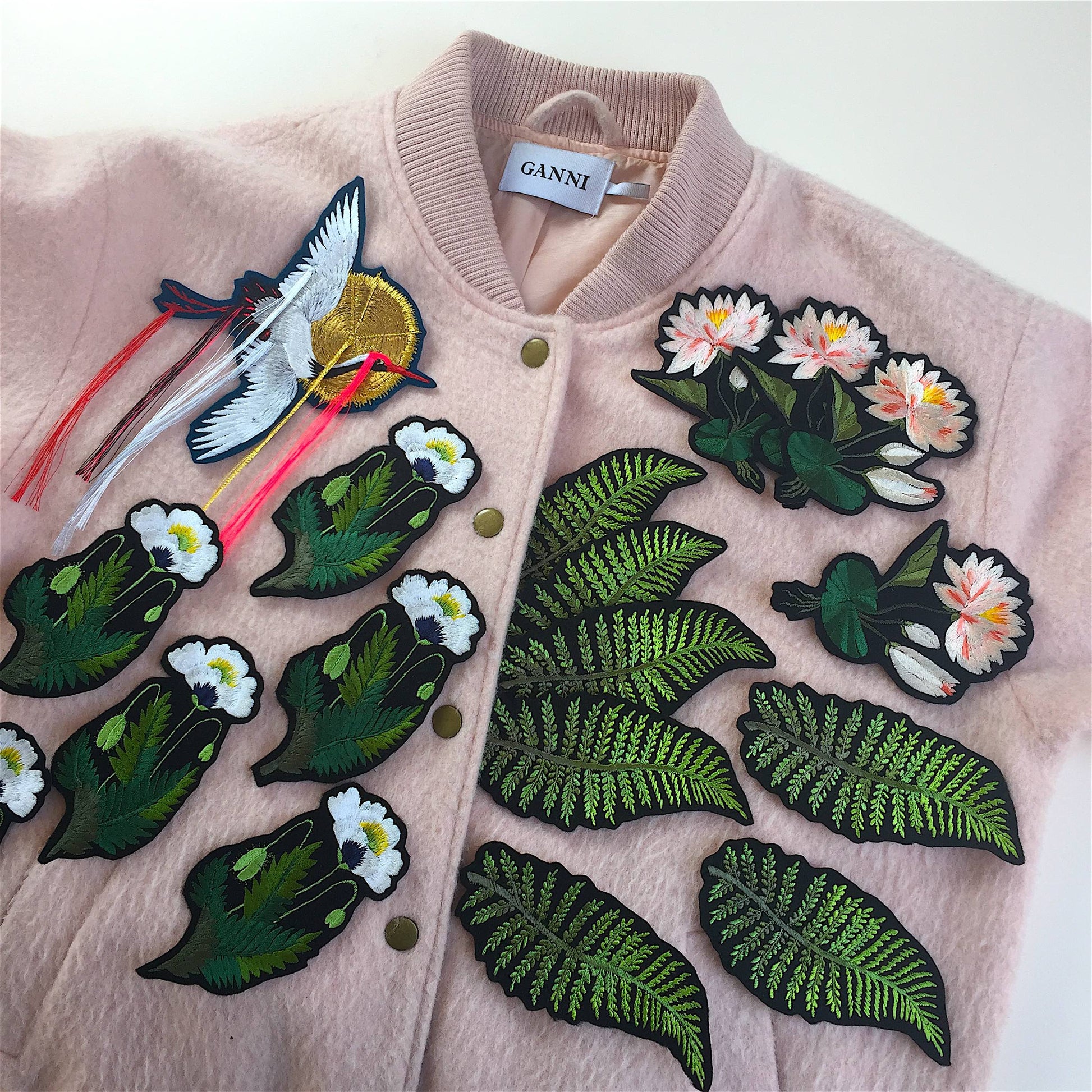 A selection of Ellie Mac embroidered patches shown scattered on a pale pink wool jacket including fringed crane, water lily, white poppy and fern